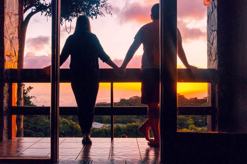 A girl and boy holding hands on a balcony, looking out at the sunset