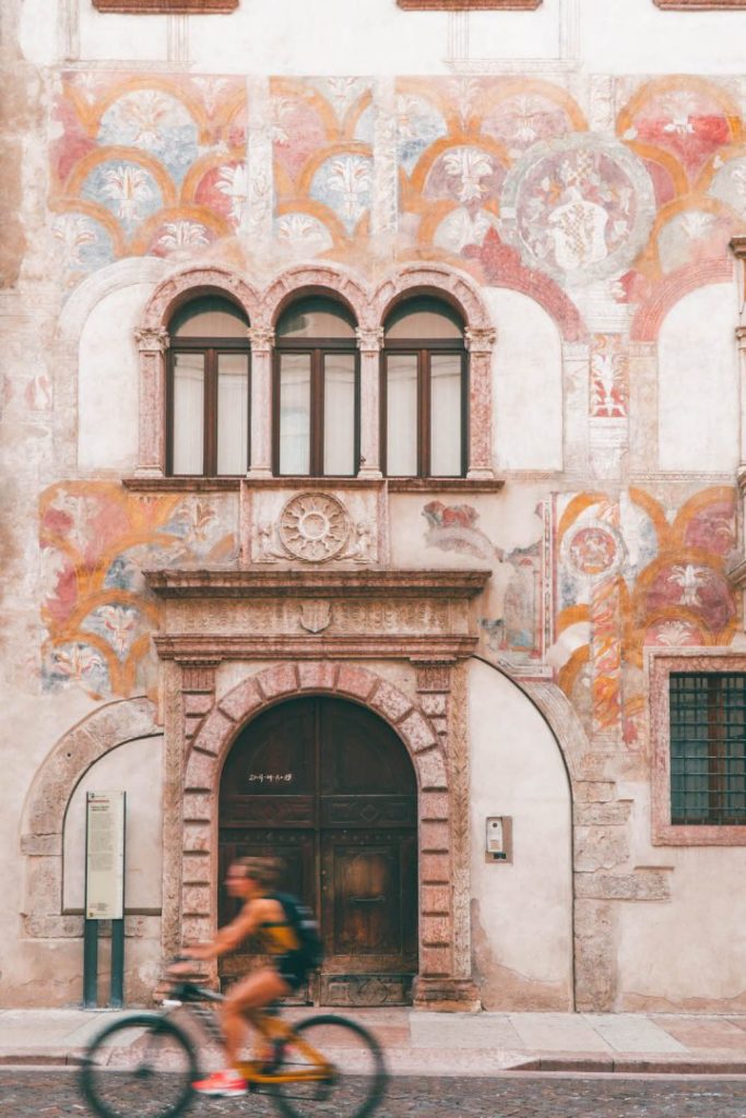A woman on a bicycle riding past a frescoed fall in Trento, Italy