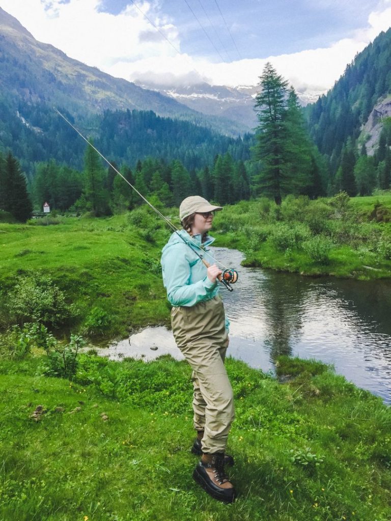 Addie in waders and holding a fishing rod in the mountains