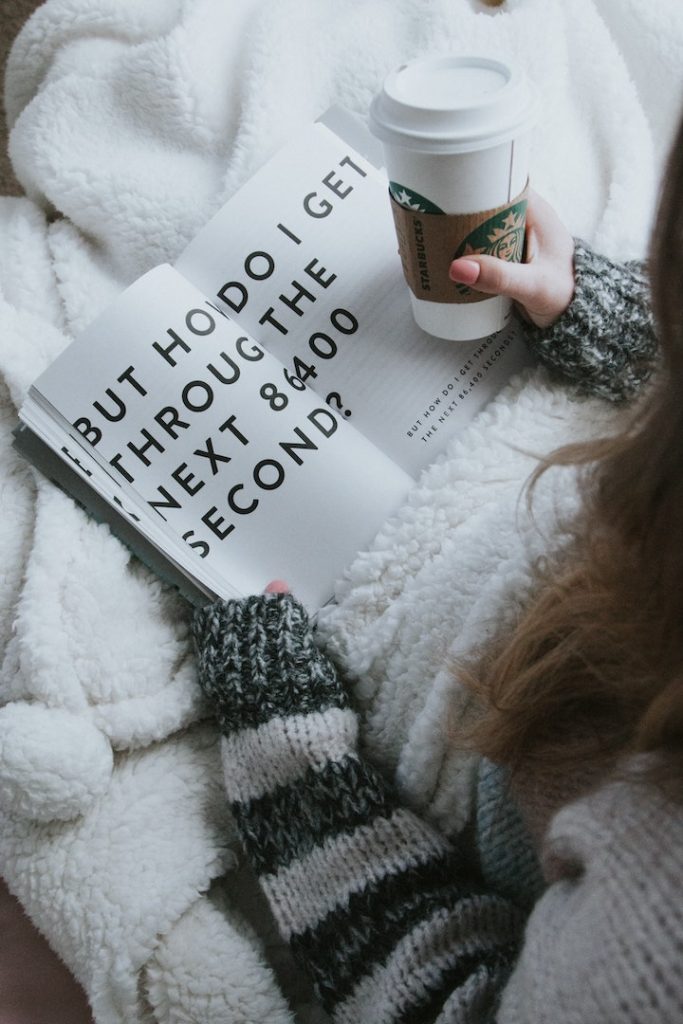 a woman sitting in bed with a fuzzy blanket, wearing a cozy sweater and holding a book and starbucks cup
