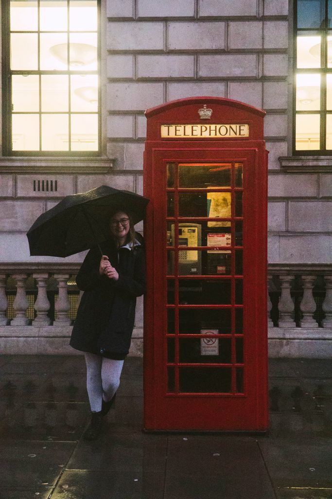 addie standing at a red phone booth holding an umbrella